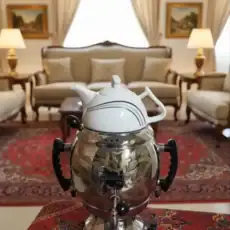 Close-up of a Khajasteh brand 6-liter Adasi samovar, a traditional Persian tea brewing device with a silver body and white porcelain top, set on a patterned red rug in a traditionally furnished room.